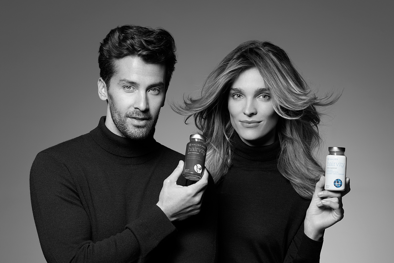 Black and white promotional image featuring a man and a woman in black turtlenecks, each holding a bottle of Hairvity hair care supplements. They both have confident, approachable smiles, and their healthy hair complements the product they're endorsing