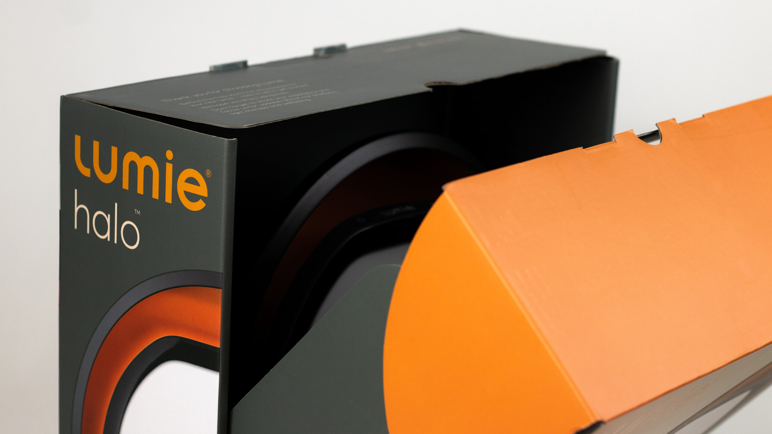 The product packaging 3D render in Unreal Engine for 'Lumie Halo', a multifunctional light therapy lamp. The box is predominantly orange and black with an image of the circular lamp on the front and the brand name 'Lumie' at the top. 